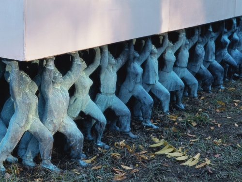 Sculpture of small figures in a row raising flat stone above their heads.