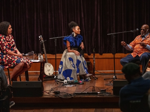 Two women and a man sit on a stage with musical instruments around them, speaking into mics.