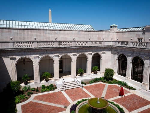 Aerial view of courtyard, surrounded by white building, with red brick pavers and a man walking in a red robe.