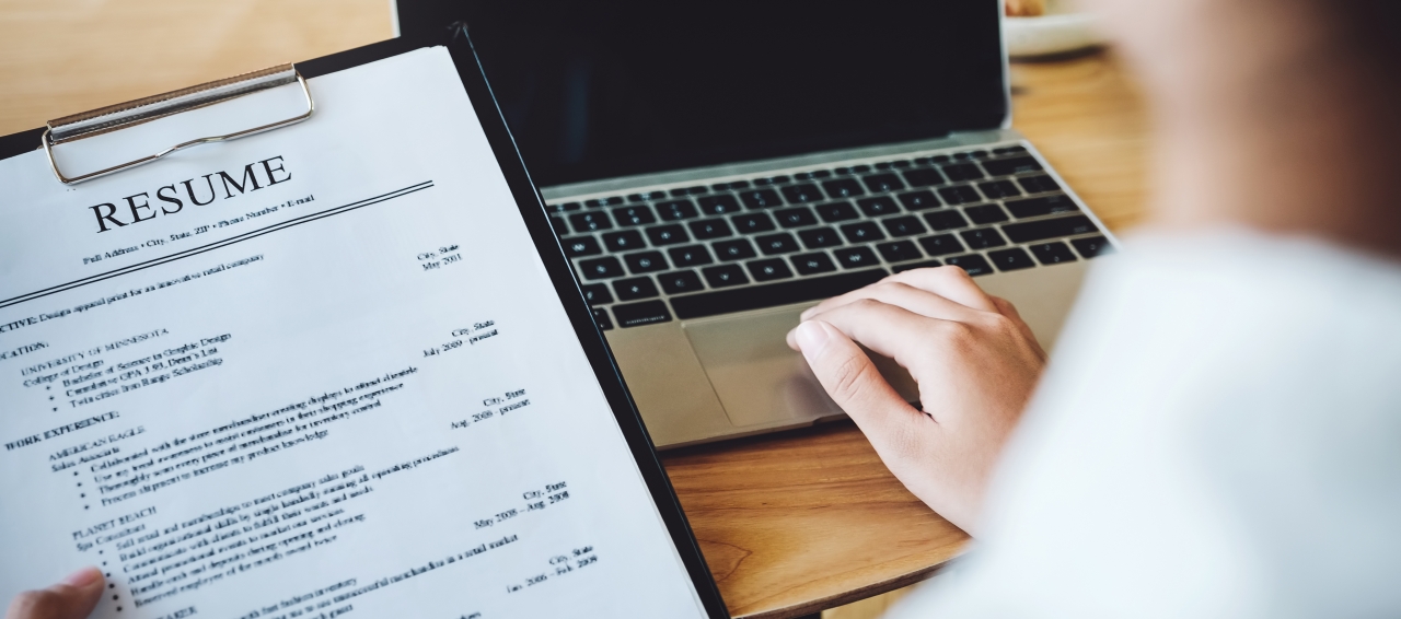 person using a laptop while holding a paper resume