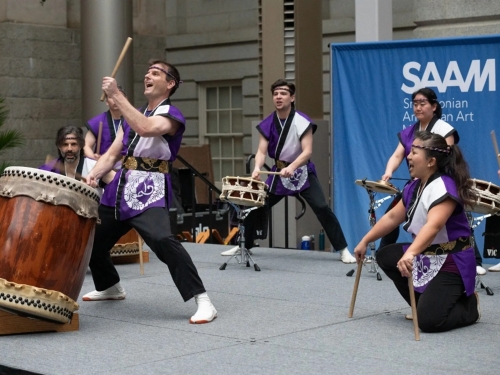 A group of people in white and purple robed tops play instruments on a small stage.