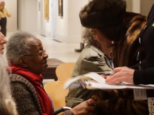 A middle-aged woman standing in front of a seated elderly woman and leans over to speak with her